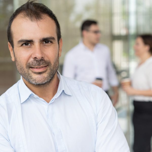 Face of happy male executive looking at camera and smiling, his colleagues talking in background. Business leader concept